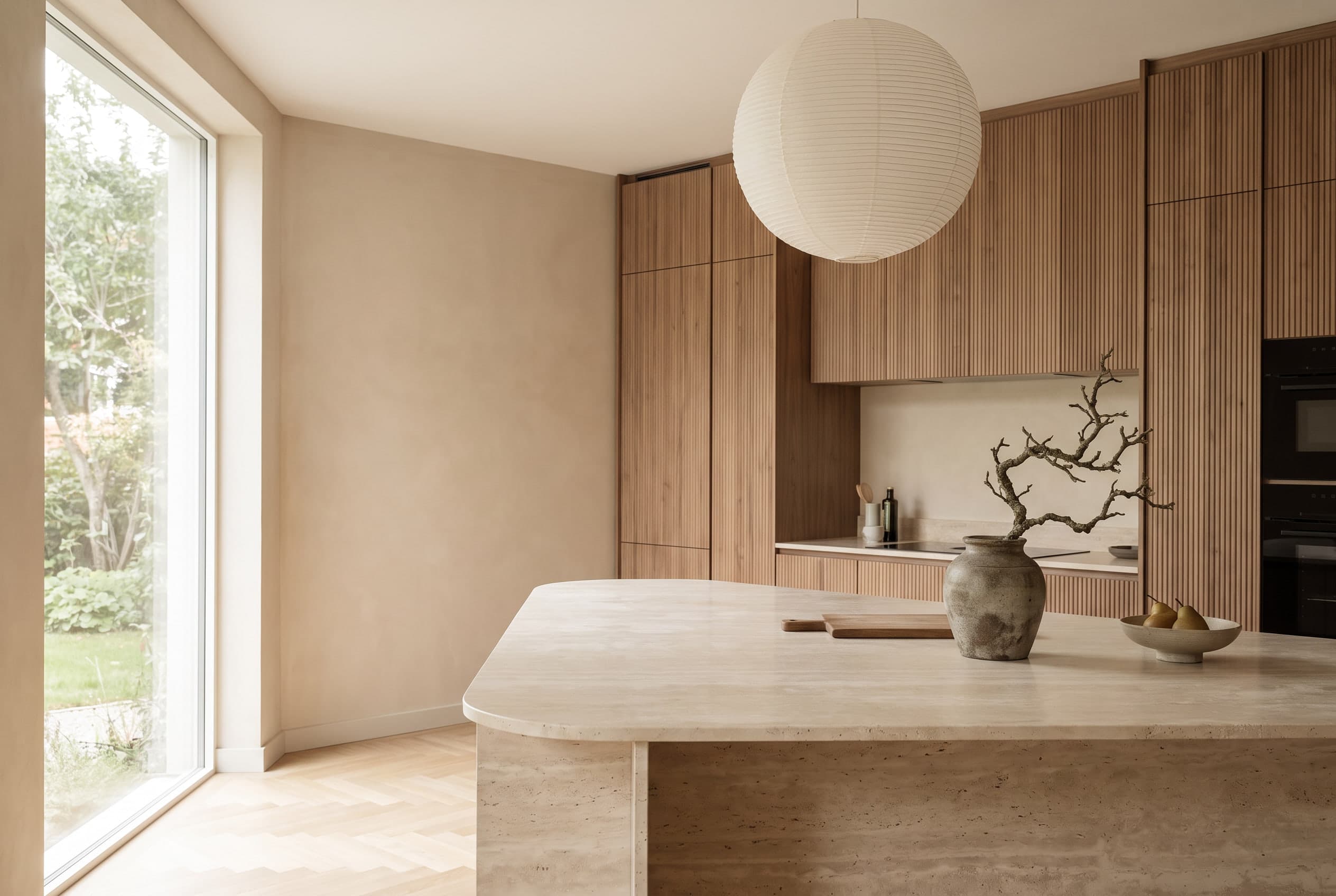 Japandi-keuken met verticaal geribbelde fronten in licht notenfineer, een warmbeige kalkgestucte wand, een eiland van travertin met afgeronde hoeken en een papieren bollamp; op het eiland een stenen vaas met een kale tak.