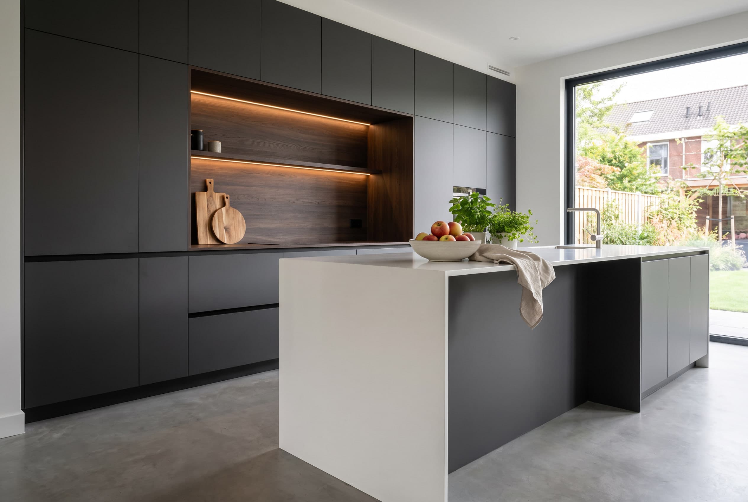 Greeploze keuken met matte antraciet fronten, een kastenwand met verlichte nis in donker houtfineer en een eiland met een wit blad dat aan de zijkant tot de vloer doorloopt, voor een groot raam met tuinzicht.