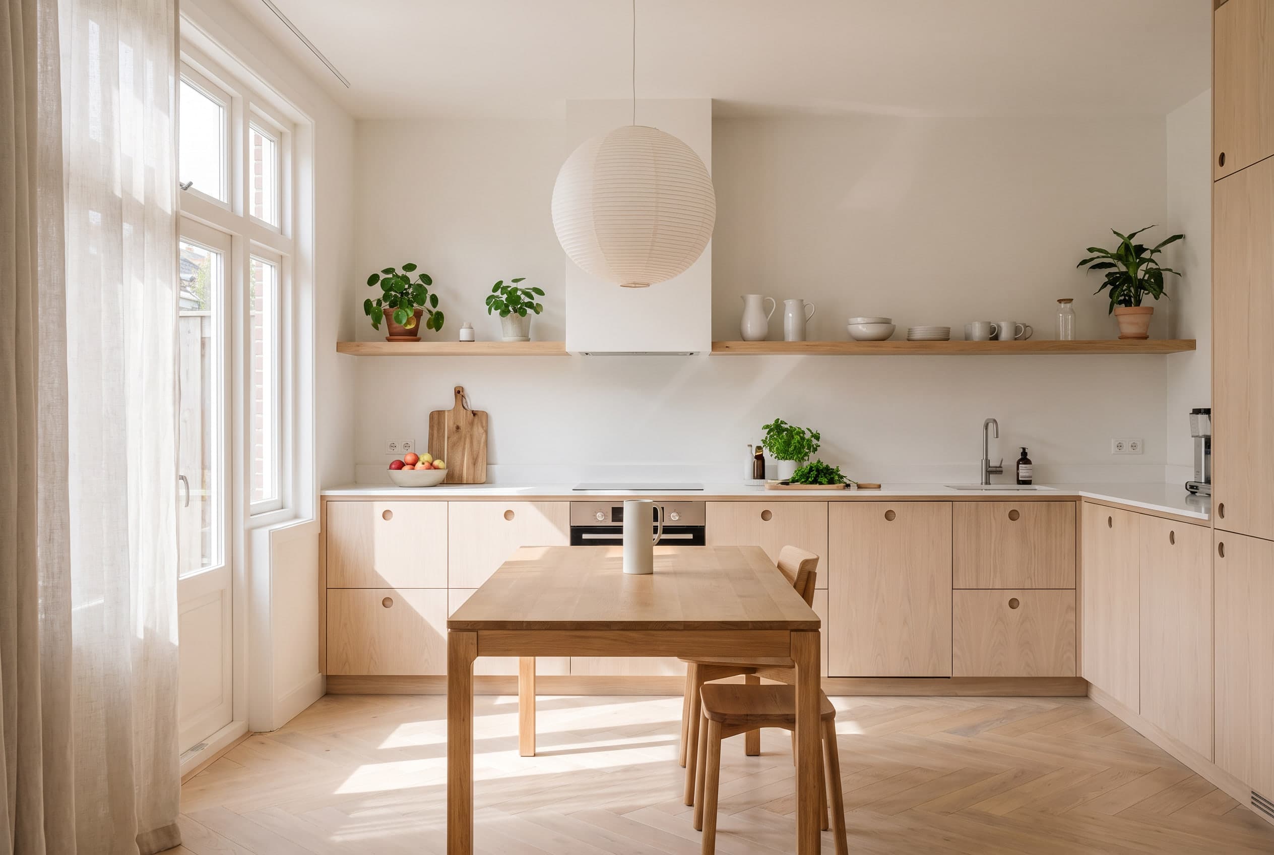 Scandinavische hoekkeuken met lichte houten fronten met ronde greepgaten, een wit werkblad, open planken met planten en wit aardewerk, een papieren bollamp en een eiken eettafel in helder daglicht.